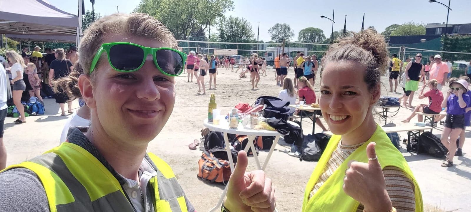 Twee lachende EHBO'ers in gele hesjes steken hun duim op bij een beachvolleybaltoernooi.