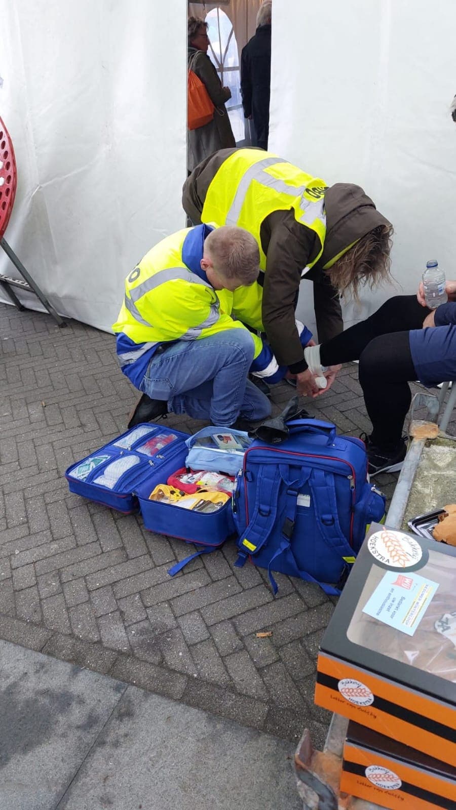 Twee EHBO'ers in gele hesjes verbinden de enkel van een bezoeker bij een tent, met een geopende EHBO-tas op de grond.