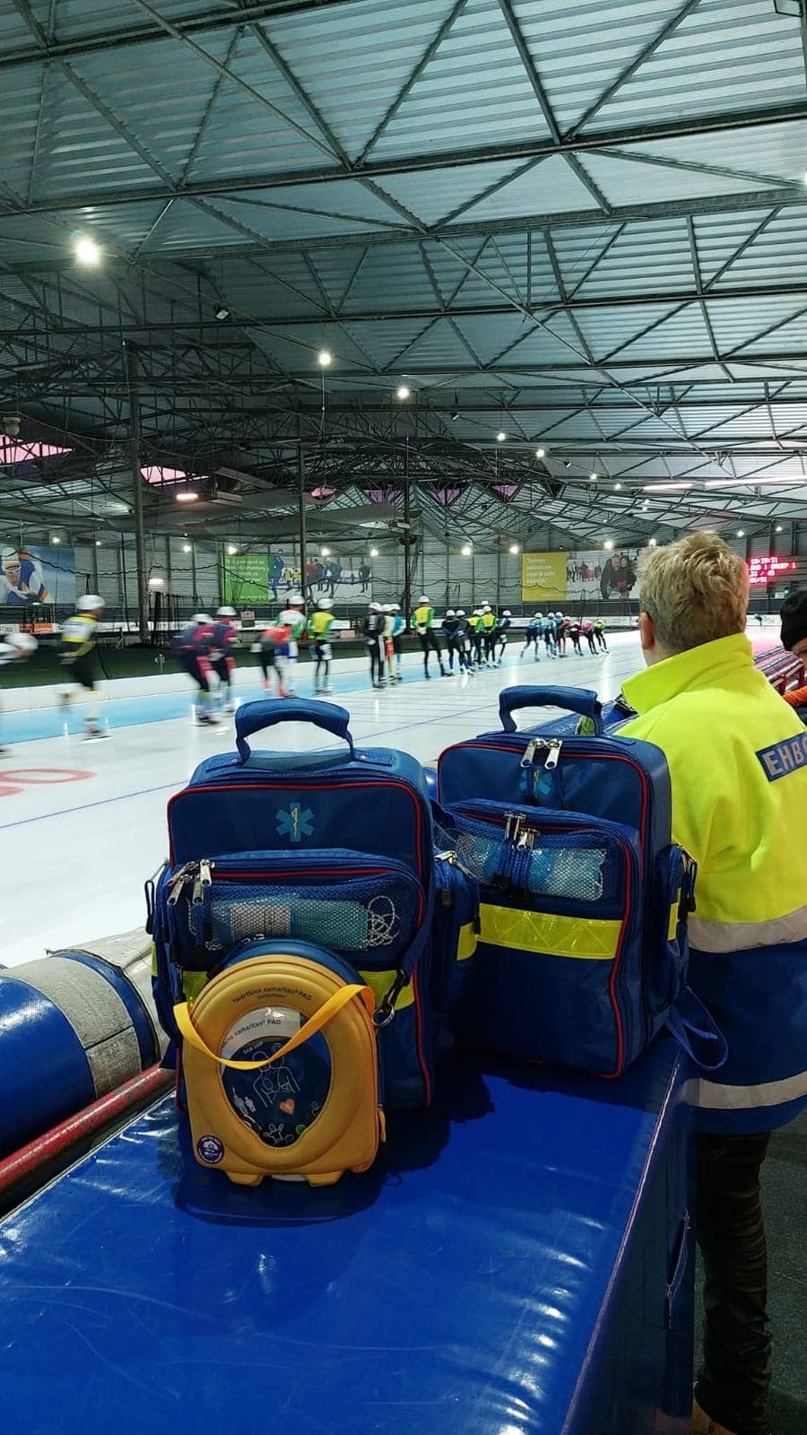 Een EHBO'er in geel jack staat naast de EHBO-tassen en AED langs de ijsbaan en kijkt naar de schaatswedstrijd.