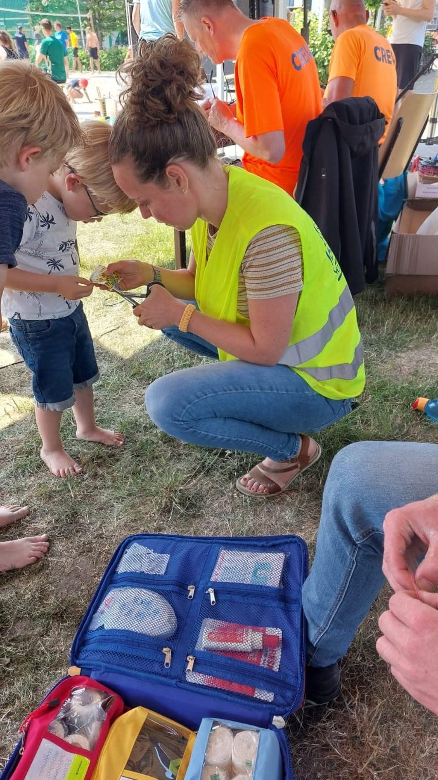 Een EHBO'er in geel hesje knipt een pleister op maat voor een jongen op blote voeten, met een geopende EHBO-tas in het gras.