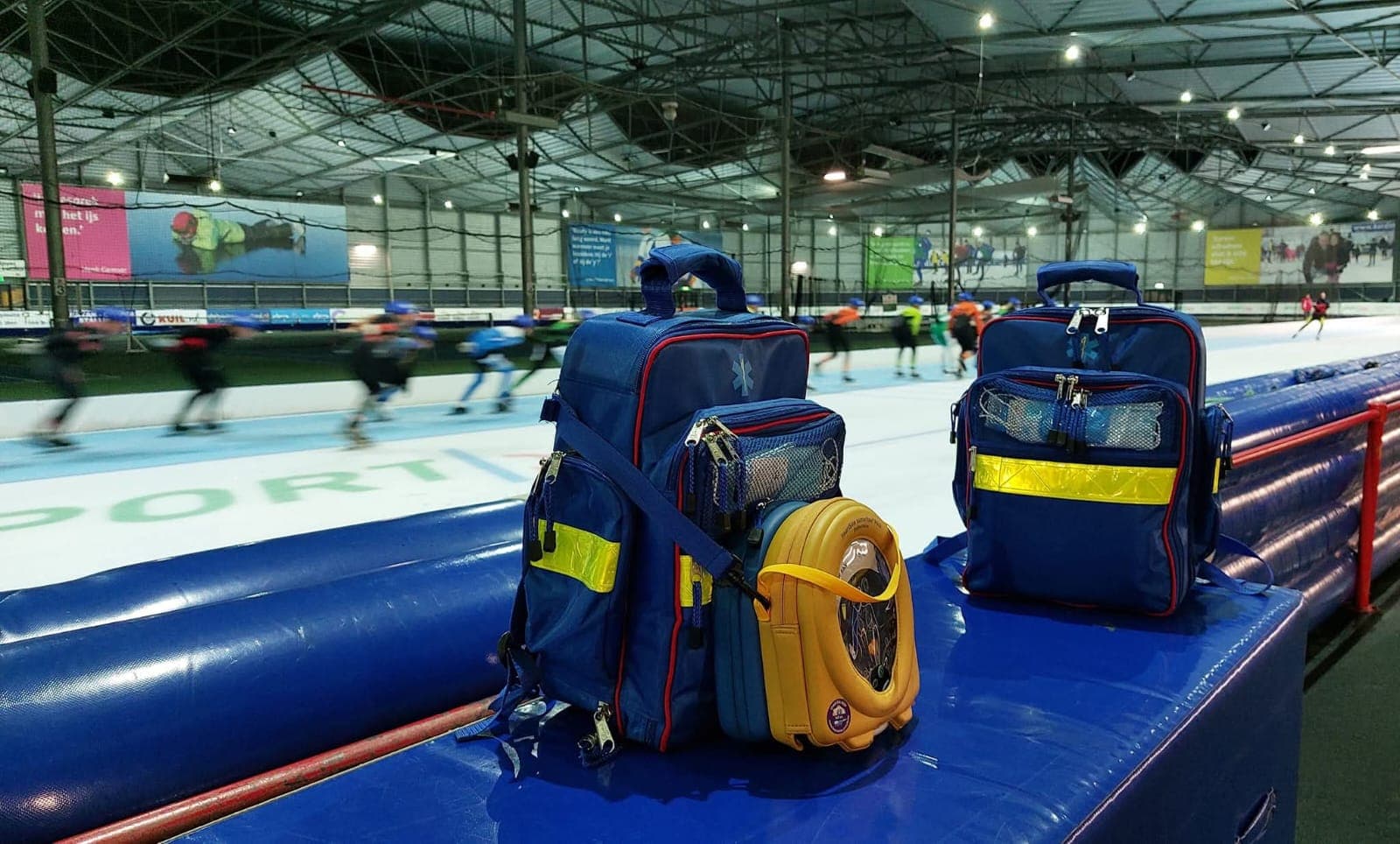 Twee blauwe EHBO-tassen en een AED staan klaar op de boarding van een ijsbaan, terwijl schaatsers voorbij komen.